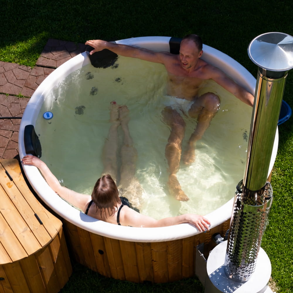 An outdoor flat bottom hot tub with two people relaxing inside, surrounded by grass and a wooden deck. A chrome chimney stack is visible on the right side.