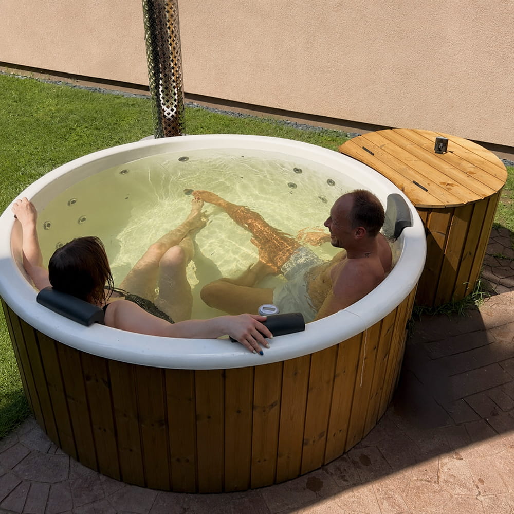 A round outdoor hot tub with a wood-look exterior and white interior, containing two people relaxing in shallow water. A matching wood-look filter box sits beside the tub on a patio, with grass and a wall in the background.