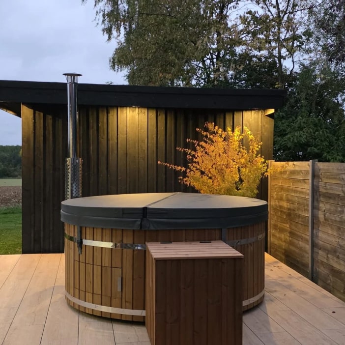 Outdoor evening view of a closed round wooden hot tub with cover near black shed and illuminated tree