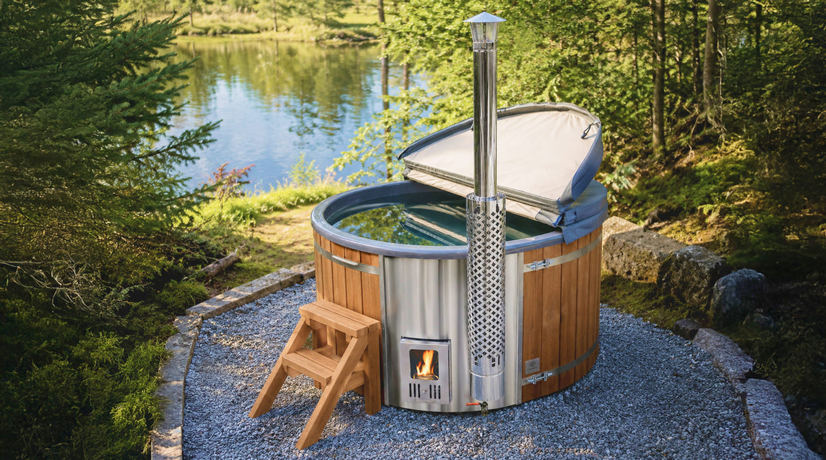 Round wood-fired hot tub with integrated stove, open cover and standard wooden steps by a forest lake