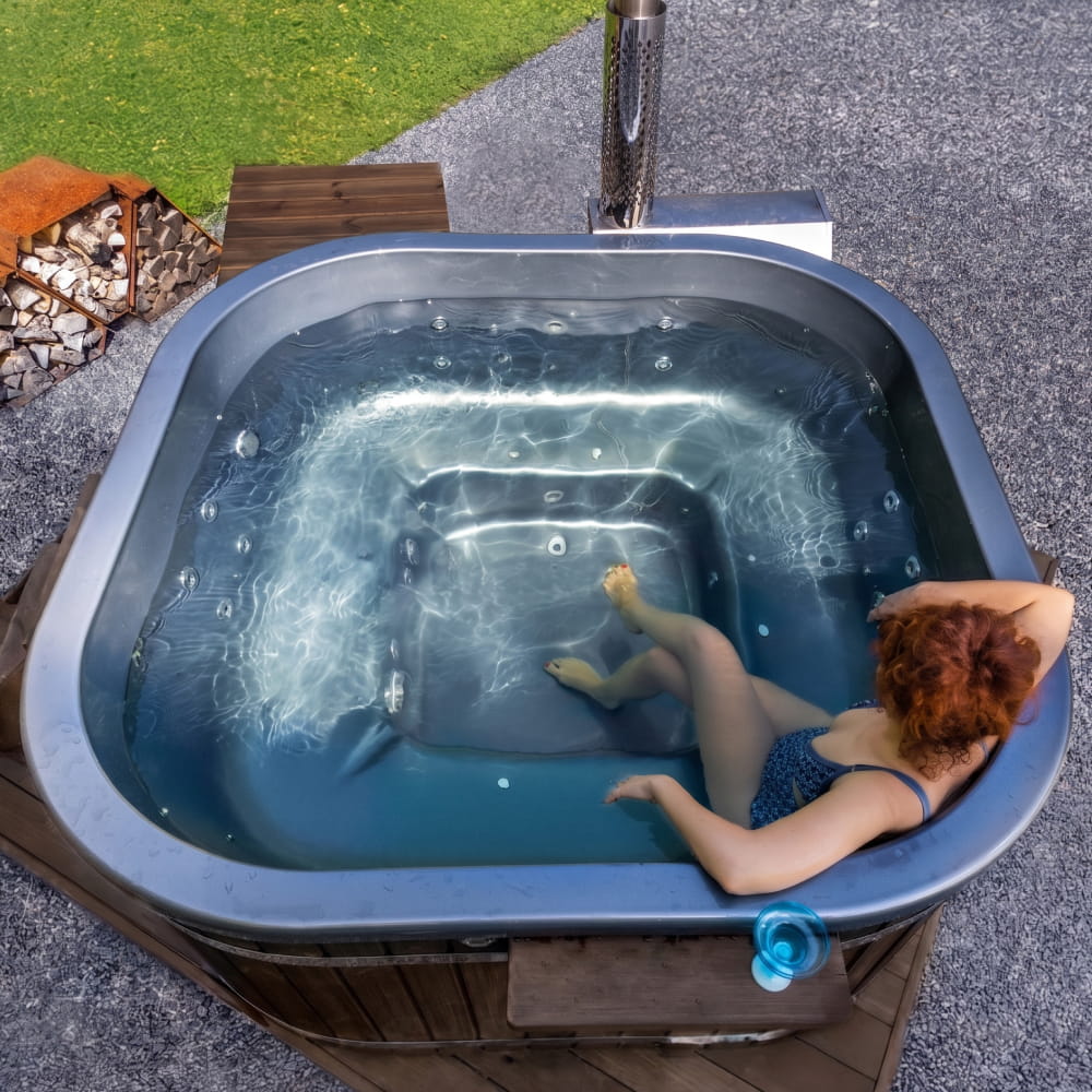 A woman relaxing in a square hot tub outdoors with a fire pit in the background