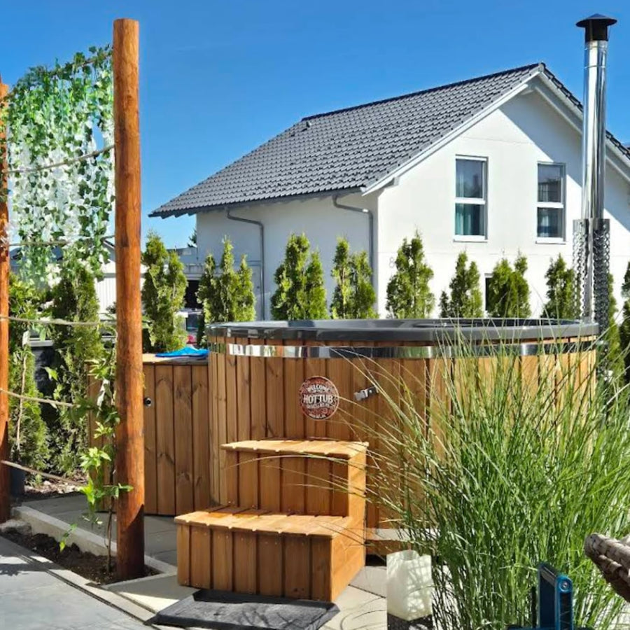 Outdoor view of wood fired hot tub with metal rim, wooden steps, and tall privacy plants beside a modern white house