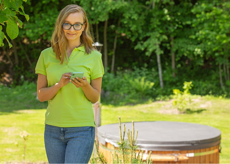 Woman in green polo shirt and glasses holding smartphone outdoors with covered wooden hot tub and green trees in background