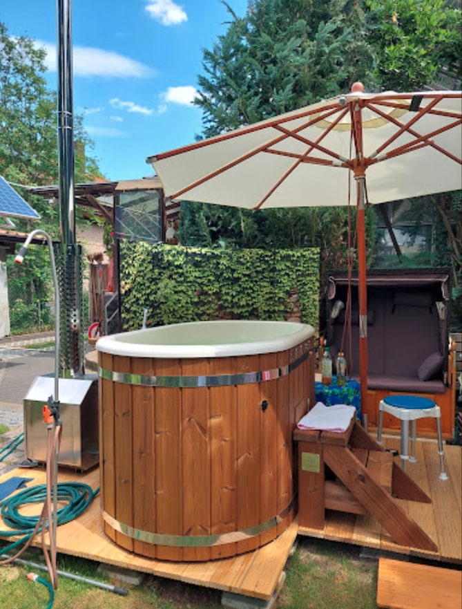 Outdoor view of a wooden hot tub with metal bands, surrounded by patio umbrella, wooden steps, and garden greenery backdrop