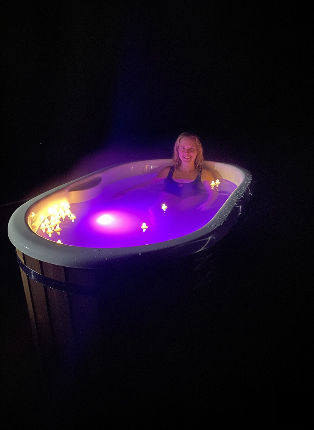 Night view of a woman relaxing in an illuminated oval hot tub with purple LED lights and floating candles