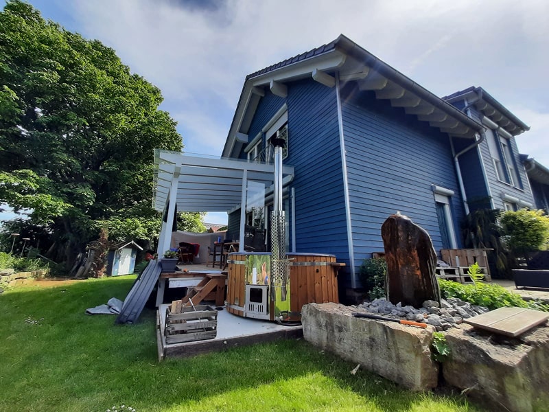 Side view of blue wooden house with covered patio, outdoor heater, firewood stack, and surrounding garden with green lawn