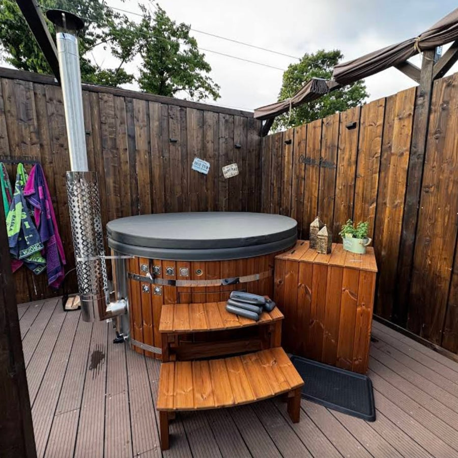 Side view of a round wood fired hot tub with closed cover, wooden steps, and privacy fence in outdoor deck area