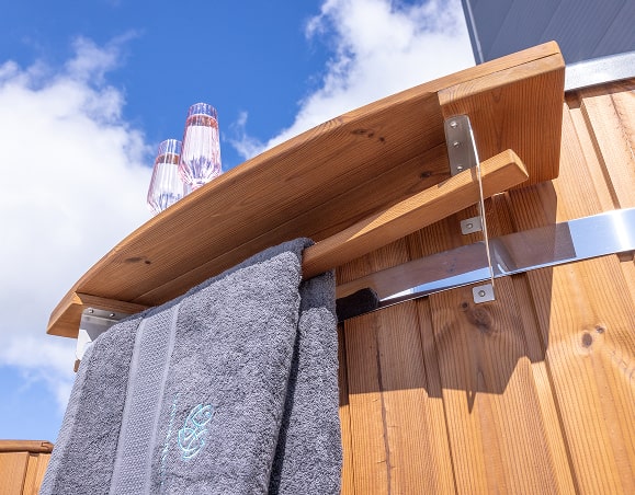 Close-up of a wooden outdoor rack attached to a wood-fired hot tub holding two gray towels under a blue sky background