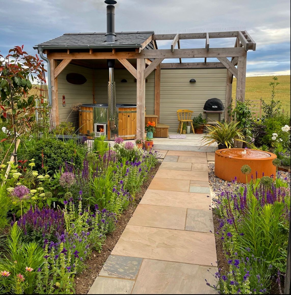 Outdoor garden view with wood pergola, hot tub, stone pathway, and flowering plants surrounding the area