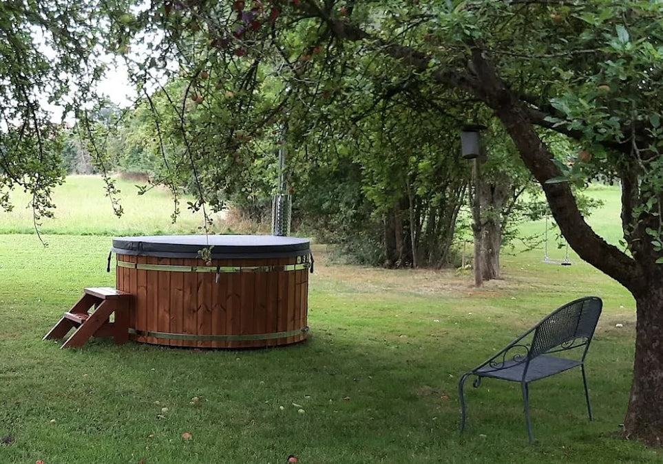 Outdoor view of wooden hot tub with cover and steps set on grassy lawn surrounded by trees and metal bench nearby