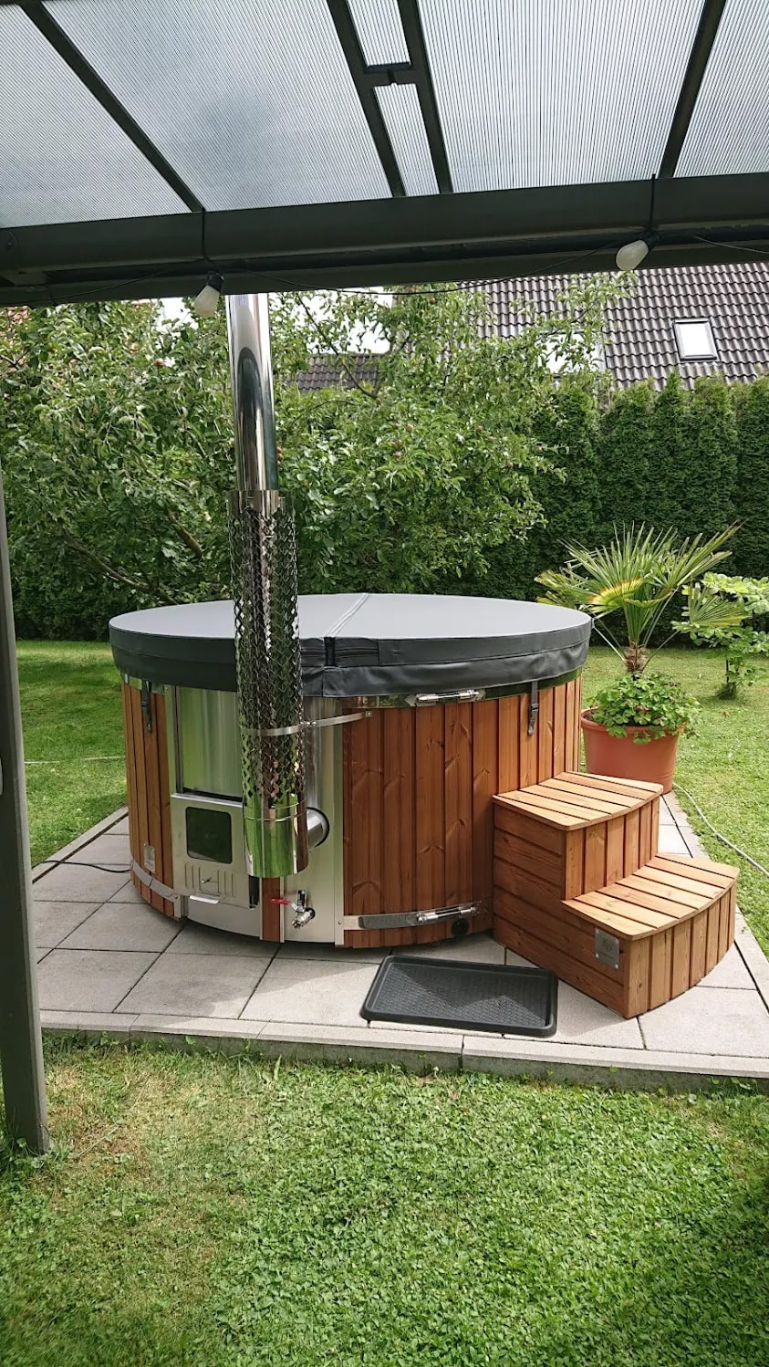 Compact Gardenvity wood fired hot tub placed under a terrace roof in a green backyard.